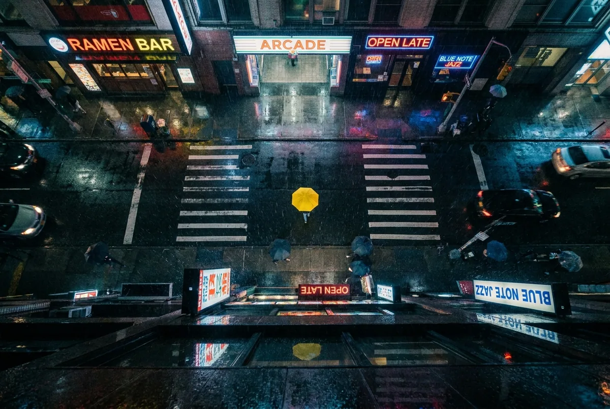Night street, aerial — Top-down aerial view of a rain-soaked city street at night, neon signs reflecting on wet asphalt, a single yellow umbrella, cinematic, moody